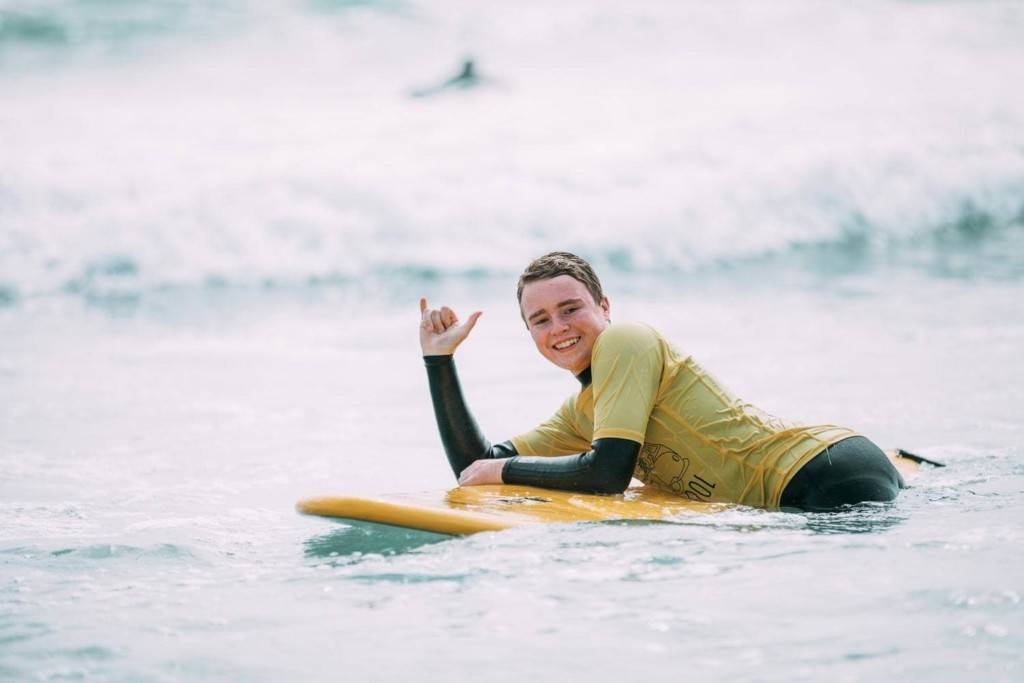 A surfing student laying down on their board in the shoreline giving the shaka sign