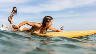 why you should go surfing, young women with long brown hair paddling on her yellow surfboard