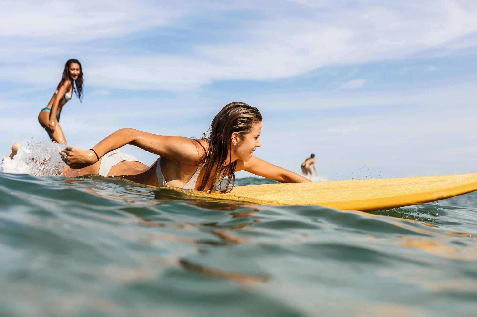 why you should go surfing, young women with long brown hair paddling on her yellow surfboard