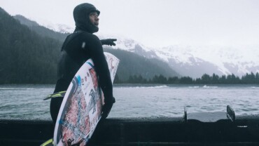 man entering water with surfboards, snowy mountain background, surfing in the winter